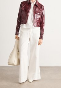 Bordowa kurtka z imitacji skóry, biała koszula w paski, białe rozkloszowane spodnie, trzyma beżową torebkę na ramię, białe szpilki z ostrym noskiem.