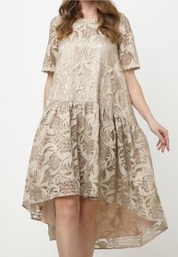 Robe en dentelle beige avec motifs floraux, manches courtes, ourlet asymétrique et jupe ample et fluide, portée par une femme de la nuque aux genoux.