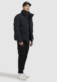 Tumesinise puffer-jakiga, millel on tikkimine ja kõrge krae. Koos mustade cargo-pükste ja valgete tossudega.