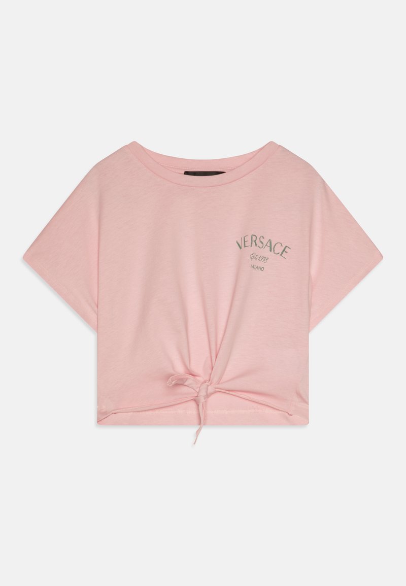 Camiseta corta de color rosa claro, hecha de algodón suave, con un detalle de lazo en el dobladillo y el logo "Versace" bordado en plata en el frente.