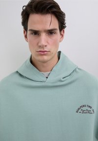 Sudadera con capucha de color verde claro y corte relajado, con un logo frontal en texto rojo oscuro. Tela suave con una textura lisa.