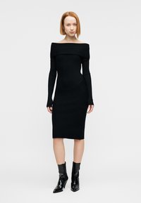 Μαύρο ριμπ πλεκτό φόρεμα με σχεδιασμό off-shoulder, μακριά μανίκια και εφαρμοστή σιλουέτα, με διαγώνια ραφή λεπτομέρεια.