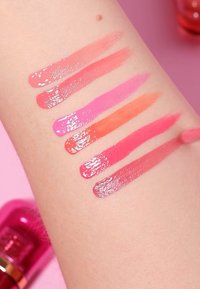 Seis muestras de productos para labios brillantes en varios tonos: durazno, rosa, coral y rojo, aplicadas sobre un tono de piel claro.