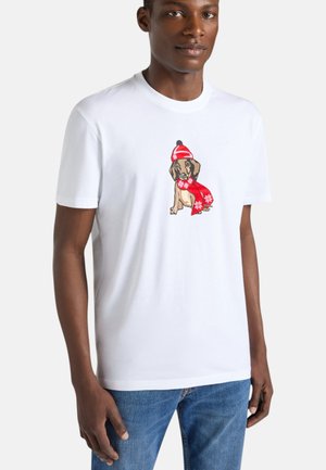 Uomo che indossa una t-shirt bianca con grafica di un cane con cappello invernale rosso e sciarpa, abbinata a jeans blu, in piedi davanti a uno sfondo semplice.