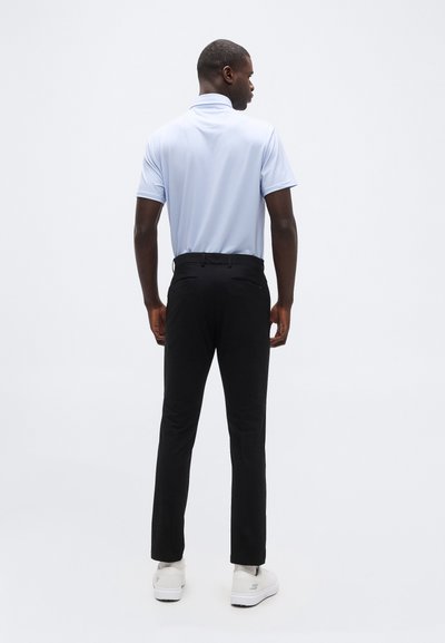 Άνδρας που στέκεται γυρισμένος με την πλάτη, φορά ανοιχτό μπλε κοντομάνικο πόλο, μαύρο παντελόνι, λευκές κάλτσες και λευκά αθλητικά παπούτσια σε απλό φόντο.