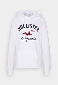 Vit luvtröja med framficka, med svart text "HOLLISTER" och en röd fågellogo, tillverkad av mjukt tyg med ribbade ärmslut.