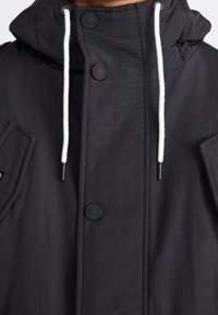 Schwarze Jacke mit Kapuze, schwarzen Knöpfen, weißem Kordelzug und zwei Vorder Taschen. Glatte Textur und minimalistisches Design.