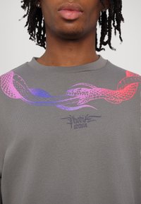Jeune personne portant un t-shirt gris avec des motifs de serpents entrelacés violets et roses sur la poitrine et le texte « Phobia archive » en dessous.