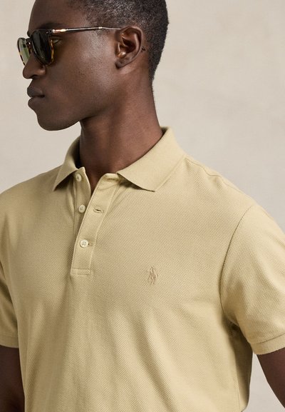 Άνδρας που φοράει γυαλιά ηλίου από κέλυφος χελώνας και μια μπεζ πικέ πόλο με μικρό κεντημένο λογότυπο, στραμμένος προς τα αριστερά σε ουδέτερο φόντο.