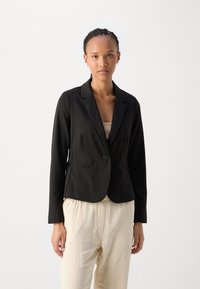 Svart figursydd blazer med en knapp, snedda slag och sidofickor. Slät tyg med en strukturerad design, bärs över krämfärgade byxor.