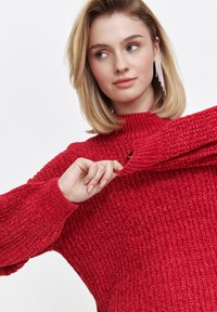 Czerwony sweter dziany z wysokim dekoltem i bufkami. Tekstura jest gruba, z prążkowanymi akcentami. Modelka częściowo unosi rękaw.
