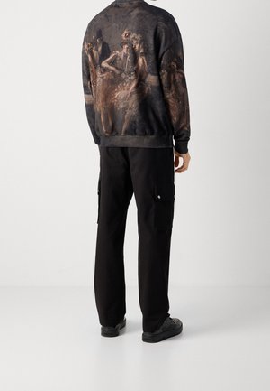 Person iført sorte cargobukser og en mørk sweatshirt med et falmet kunstnerisk tryk af balletdansere på ryggen, stående mod en ensartet baggrund.