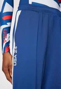Modré športové nohavice s bielymi bočnými akcentmi, nápisom "USA 24" vytlačeným v bielej farbe na nohe a hladkou, štruktúrovanou tkaninou.