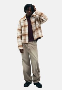Hnedo-krémová károvaná bunda s golierom, oblečená cez vínovú košeľu, skombinovaná s béžovými nohavicami s širokými nohavicami a čiernymi platformovými topánkami.