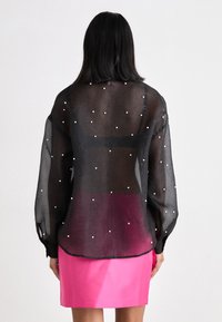 Schwarze transparente Bluse mit langen Ärmeln, geschmückt mit weißen Punkten. Getragen über einem pinken Rock mit rundem Saum und leichter Transparenz.