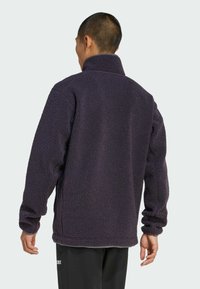 Veste en polaire violet foncé avec un col montant, un tissu texturé, et des manches longues, offrant une coupe décontractée et des poignets côtelés.