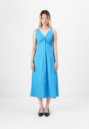 Blaues ärmelloses Kleid mit V-Ausschnitt, verziert mit Falten und drei dekorativen Knöpfen an der Brust; kombiniert mit schwarzen spitzen Schuhen.