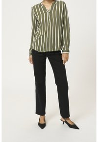 Femme portant un haut olive avec des rayures verticales blanches, un pantalon noir à jambes droites et des escarpins noirs à bout pointu, se tenant debout.