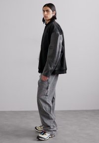 Jachetă oversized neagră cu mâneci din piele, combinată cu pantaloni gri largi cu fermoare și adidași colorați cu talpă groasă.