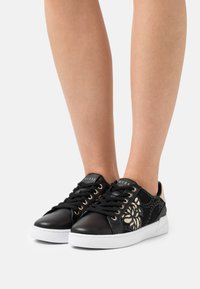 Baskets noires avec un bout en cuir lisse, des côtés en mesh, des détails floraux dorés, une semelle blanche et des lacets noirs. Comprend un logo sur la languette.