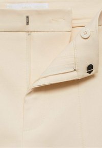 Pantalons couleur crème en tissu lisse, avec une fermeture éclair sur le côté, un bouton à l'avant et une taille cintrée ornée de coutures.