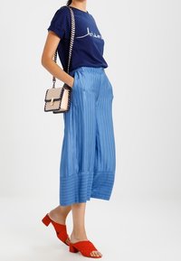 Žena v námořnickém modrém tričku, širokých světle modrých pruhovaných kalhotách, červených sandálech na podpatku a s béžovou kabelkou na řetízkovém popruhu.