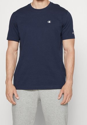 Hombre que lleva una camiseta de manga corta azul marino de Champion con pequeños logotipos en el pecho y la manga, combinada con pantalones deportivos de color gris claro.