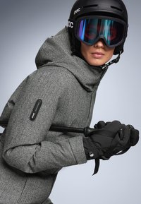 Sivá lyžiarska bunda v rybej kostre s kapucňou, v kombinácii s čiernymi rukavicami a prilbou. Vyznačuje sa štýlovým dizajnom a integrovaným vybavením.