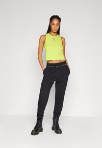 Neon groene cropped tanktop met logo, gecombineerd met zwarte joggerbroeken met trekkoord. Model draagt zwarte veterschoenen. Effen achtergrond.