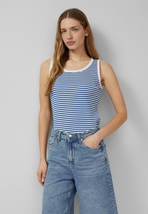 Femme aux longs cheveux portant un haut sans manches rayé bleu et blanc et un jean bleu ample taille haute, une main dans la poche, sur fond gris.