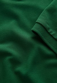 Tissu vert texturé avec armure visible et détail de revers plié, suggérant une partie d’un vêtement comme une chemise ou un pull.