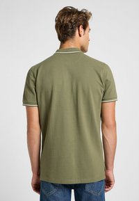 Oliivivihreä lyhythihainen polo-paita, jossa on yksinkertainen kaulus ja valkoiset yksityiskohdat hihansuiden reunassa. Sileä puuvillainen pinta.
