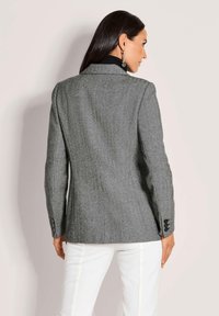 Grijze herringbone blazer met een getailleerde pasvorm, notch-kraag en zwarte afwerking. Voorzien van een vierknopen sluiting bij de manchetten en een gladde textuur.