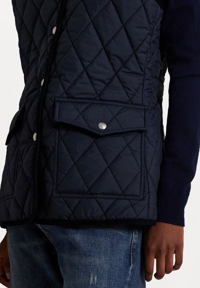 Marineblauwe gewatteerde vest met een diamanten patroon, met een voorkrui pocket met drukknoopsluiting en een afgeronde zoom. Gekoord met blauwe denim jeans.