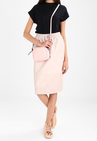 Donna che indossa una maglietta nera, una gonna midi rosa chiaro, una borsa a tracolla rosa chiaro e scarpe da ginnastica metalliche color oro rosa, in piedi con le gambe incrociate.