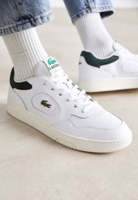 Biele kožené tenisky s zelenými akcentmi, okrúly špička, perforované detaily a textilná nášivka s logom Lacoste na jazyku.