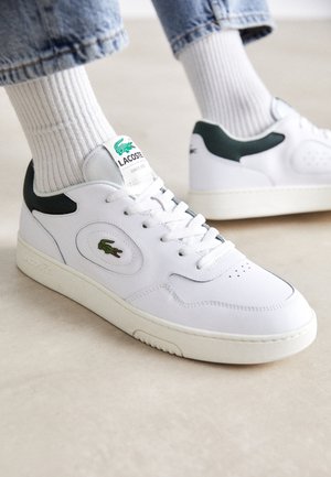 Zapatillas de cuero blanco con acentos verdes, punta redonda, detalles perforados y una etiqueta de tela con el logotipo de Lacoste en la lengüeta.