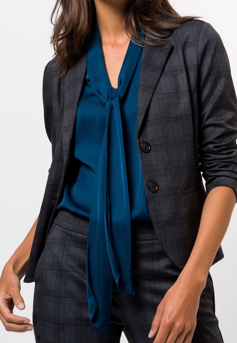 Blazer de cuadros gris con dos botones, combinado con una blusa de satén color teal con un cuello anudado. Tela suave y ajuste entallado.