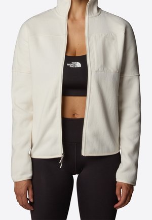 Kvinde iført en cremefarvet fleecejakke med lynlås over en sort sports-bh og leggings, begge med The North Face-logo synlig.