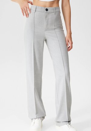Femme portant un pantalon large taille haute gris clair avec des plis à l'avant et une fermeture à boutons, associé à des baskets blanches.