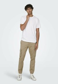 T-shirt blanc à col rond associé à un pantalon cargo beige doté de poches latérales, de poignets élastiques et de chaussures de sport blanches. Design simple et fonctionnel.