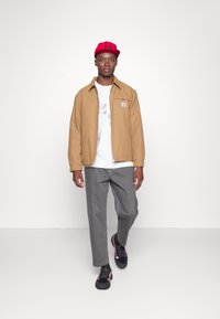 Chaqueta ligera marrón, camiseta gráfica blanca, pantalones grises y zapatillas negras con detalles rojos. Gorra roja con el logo de "Carhartt".