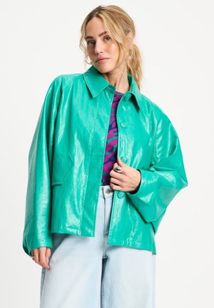 Vrouw met blond haar in een staart draagt een glanzend teal gekleurd jasje met knopen over een paarse top met patroon en lichtblauwe wijde jeans.