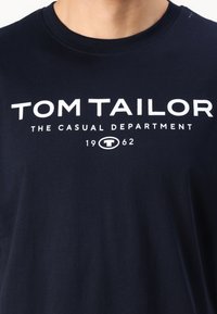Tamsiai mėlynas medvilninis marškinėlis su baltu tekstu, kuriame rašoma "TOM TAILOR THE CASUAL DEPARTMENT 1962" moderniu, ryškiu šriftu.