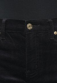 A fekete kordbársony nadrág karcsúsított szabású, arany színű fémrészekkel és varrási mintázattal a zsebek és a derékrész előtt.