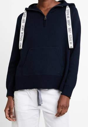 Osoba ubrana w granatową bluzę z kapturem na pół zamka, z białymi sznurkami i napisem, w połączeniu z białymi spodniami i paskiem w paski.