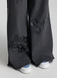 Pantaloni larghi in denim nero con orli sfrangiati, stampe di testo nere, indossati sopra scarpe bianche con calzini blu.
