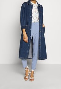 Langer Jeansmantel mit kragenähnlichem Ausschnitt, Frontknöpfen und zwei aufgesetzten Taschen, getragen über einem blumigen Hemd und hellblauen Hosen. Weiße Riemchensandalen.