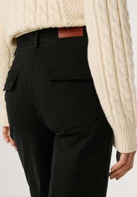 Zwarte hoge cargobroek met twee achterkantzakken, gecombineerd met een crèmekleurige geribbelde sweater. Bevat een bruine leren merklabel op de taille.