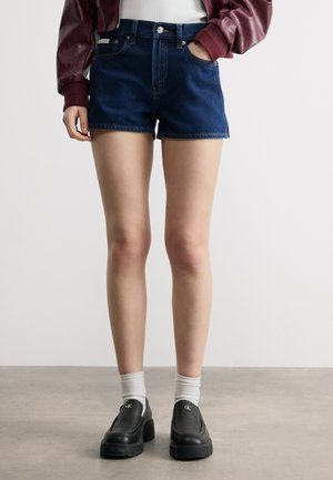 Άτομο που φοράει σκούρα μπλε τζιν σορτς, μαρόν μπουφάν, λευκές κάλτσες και μαύρα παπούτσια χωρίς κορδόνια σε απλό φόντο.